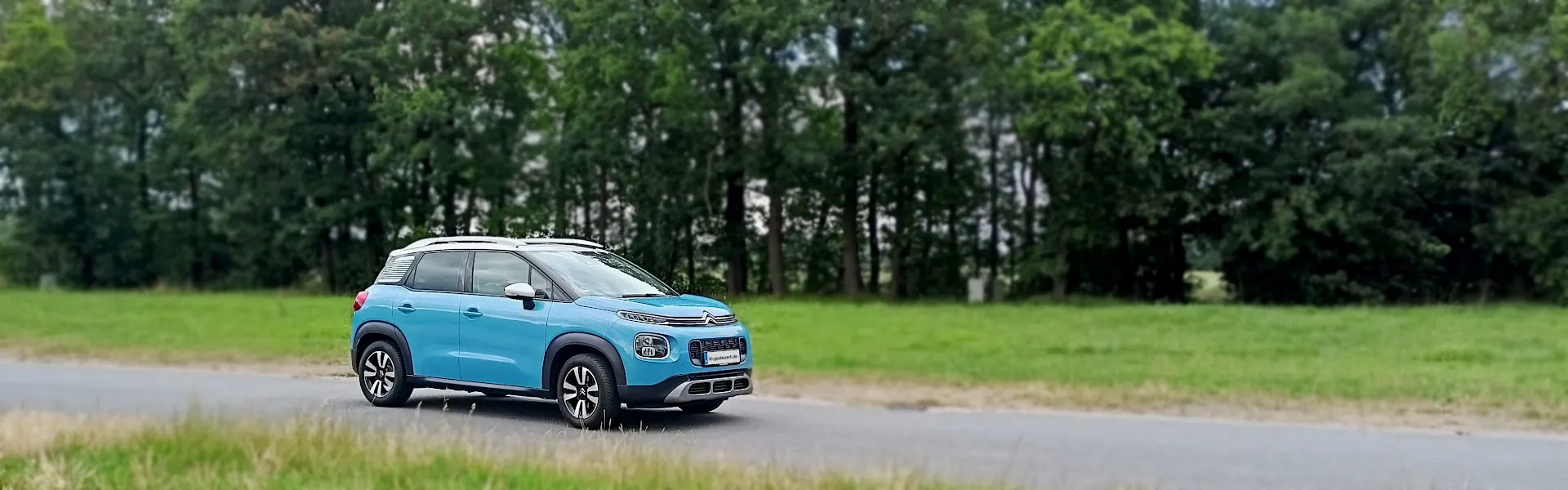 Bild 1: Ki-gesteuerter hellblauer Citroen C3 Aircross mit weißem Dach fährt auf einer Strasse vor Bäumen