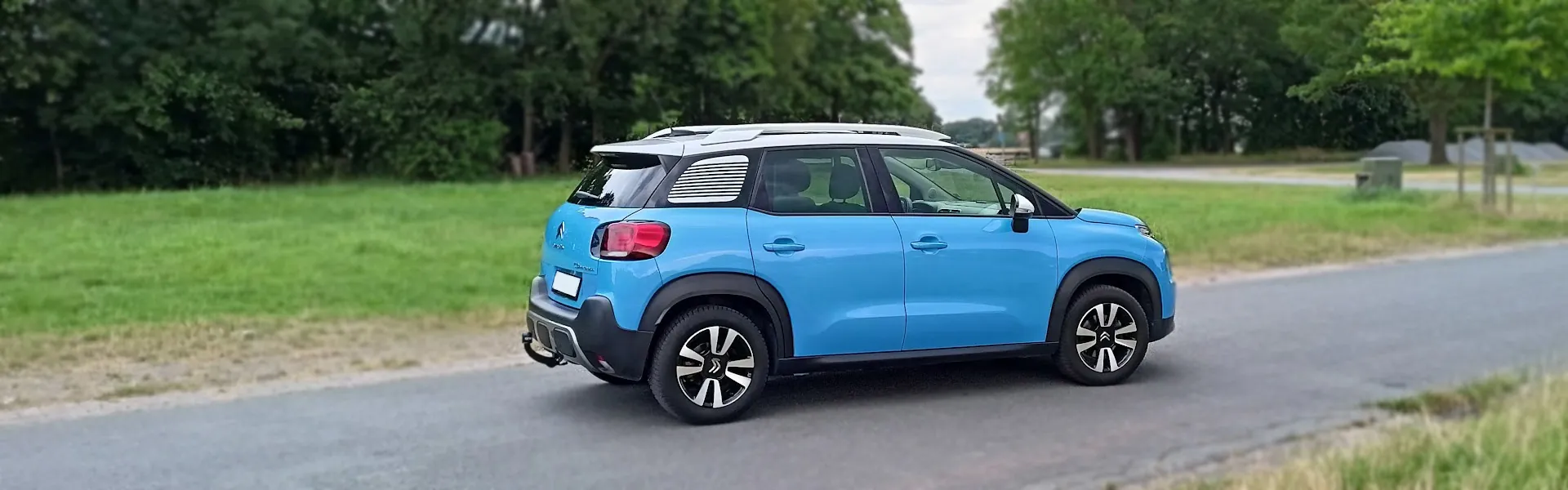 Bild 2: Ki-gesteuerter hellblauer Citroen C3 Aircross mit weißem Dach fährt auf einer Strasse vor Bäumen